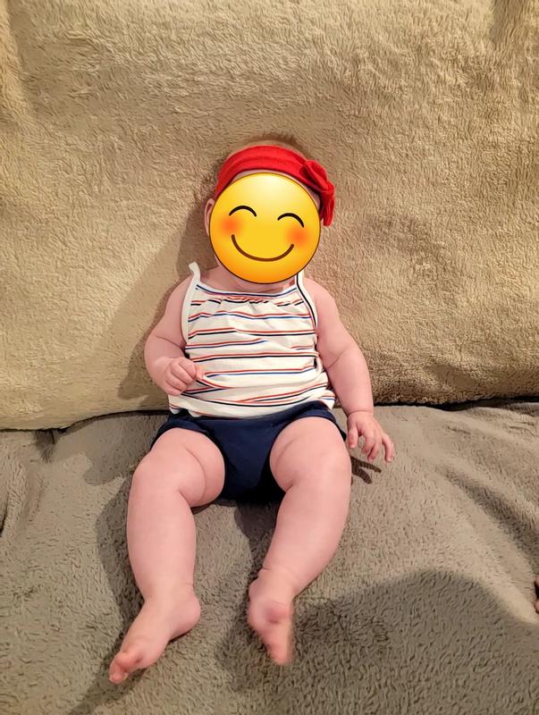 Smiley face infant in a red, white, and blue striped tank with navy blue bloomers and a red headband sitting on a couch.