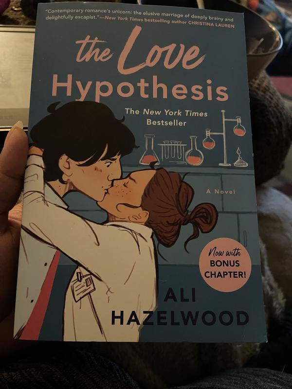 Image ID: Black person’s hand holding Ali Hazelwood’s The Love Hypothesis which features a blue cover with a tall Caucasian man wide-eyed in surprise with prominent nose. He is being kissed by a Caucasian woman with brown hair and her eyes closed.