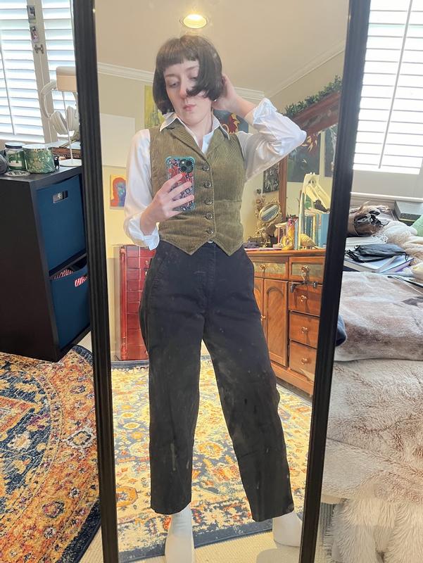 The JNY button-up is neatly tucked into black high-waisted jeans. The collar of the button-up folds over a v-neck cord buttoned vest in olive green. The sleeves of the button-up are cuffed at the wrist. The pits of the top are unobstructed by the vest.