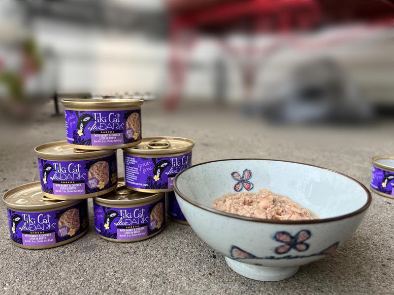 Bowl with wet cat food in it sits next to six 3oz cans of Tiki Cat After Dark: Shreds With Rabbit and Chicken Liver in Broth.  They are stacked in a pyramid.