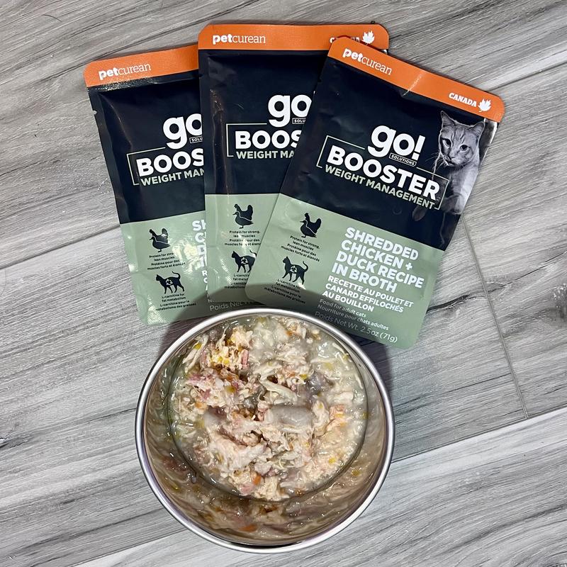 Three packets of Go! Solutions Booster Weight Management wet food in the flavour Shredded Chicken + Duck Recipe In Broth are laid around a bowl of the same food.